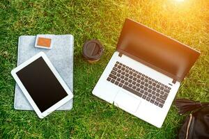 Laptop computer on green grass with coffee cup, bag and tablet in outdoor park photo