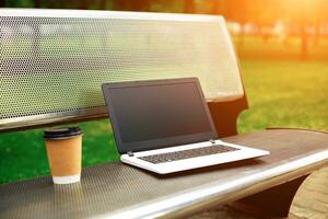 Mockup image of laptop with blank black screen and coffee cup on metal bench in nature outdoor park photo