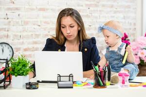 Cheerful young beautiful businesswoman looking at laptop while sitting at her working place with her little daughter photo