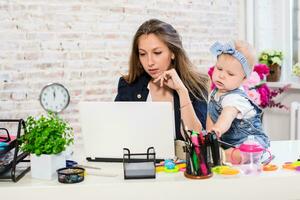 alegre joven hermosa mujer de negocios mirando a ordenador portátil mientras sentado a su trabajando sitio con su pequeño hija foto