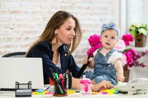 Cheerful young beautiful businesswoman looking at telephone while sitting at her working place with her little daughter photo