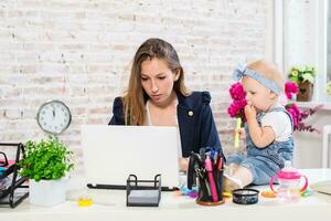 alegre joven hermosa mujer de negocios mirando a ordenador portátil mientras sentado a su trabajando sitio con su pequeño hija foto