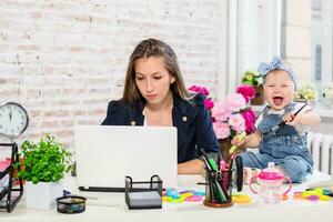 Cheerful young beautiful businesswoman looking at laptop while sitting at her working place with her little daughter photo