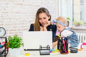 alegre joven hermosa mujer de negocios mirando a ordenador portátil mientras sentado a su trabajando sitio con su pequeño hija foto