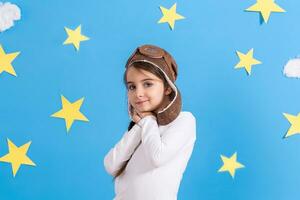 pequeño contento niña en de aviador gorra sonriente y jugando, en blanco camisa a el estudio. azul cielo, blanco nubes y estrellas antecedentes. foto