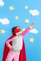 Little child plays superhero. Kid on the background of bright blue wall with white clouds and stars . Girl power concept. photo