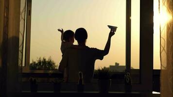 Mom holds her daughter in her arms standing at the window and launches a paper plane during sunset. video