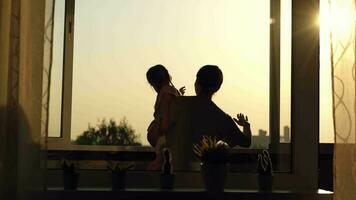 alegre mamá y hija son jugando imitando el vuelo de un avión mientras en pie cerca el ventana durante puesta de sol. video