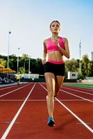 Sporty fitness woman jogging on red running track in stadium. Training summer outdoors on running track line with green trees on background. photo