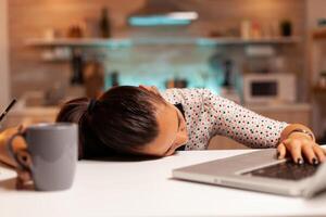 Freelance woman falling asleep in home kitchen working overtime on a project. Employee using modern technology at midnight doing overtime for job, business, busy, career, network, lifestyle ,wireless. photo