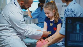 Little girl having arm bandaged sitting in bed during consultation. Healthcare physician specialist in medicine providing health care service radiographic treatment examination in hospital cabinet photo