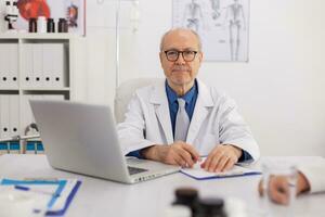 Portrait of senior man specialist physician looking into camera sitting at desk in meeting room working at medical expertise. Doctor prescribing pill medication treatment writing on clipboard photo