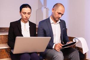 Overworked tired businessman doing overtime sitting on stairs helping coworker with diffcult deadline. Colleague entrepreneur working late together in evening at corporate job sitting on staircase, explaining difficult finance project. photo