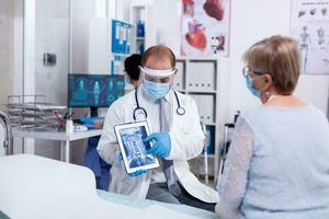 Doctor in hospital room explaining xray on tablet for senior woman during consultation in clinic room. Medical examination for infections, disease and diagnosis. photo