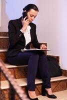 Overworked businessoman disccusing about project deadling on smartphone late at night doring overtime. Serious entrepreneur working on job project sitting on staircase of business building at night hours for job. photo