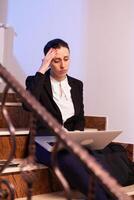 Exhausted worried businesswoman using laptop to finish corporate project deadline. Serious entrepreneur working on job project sitting on staircase of business building at night hours for job. photo