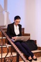 Stressed exhausted businesswoman working on to finish a business job deadline at late hours. Serious entrepreneur working on job project sitting on staircase of business building at night hours for job. photo