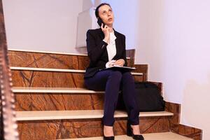 Stressed exhausted businesswoman having a phone conversation about project deadling during overtime. Serious entrepreneur working on job project sitting on staircase of business building at night hours for job. photo