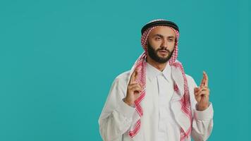 Muslim man holds fingers crossed and praying to allah over blue background, showcasing religion and spiritual belief. Young adult in islamic clothes being religious, worshipping God with devotion. photo