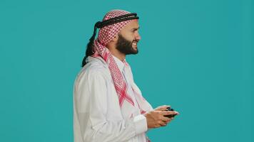 Modern person playing online games with controller, having fun with video gaming tournament over blue background. Young muslim gamer in national costume plays competition in studio. photo