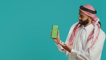 Middle eastern man points at greenscreen display on smartphone, showcasing isolated copyspace template on mobile device screen. Young person in islamic costume shows blank chromakey. photo