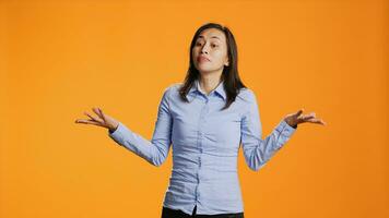 Clueless person raising shoulders on camera, expressing doubt and being uncertain about answer. Young asian woman acting unsure and confused standing against orange background. photo