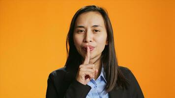 Filipino person shows hush gesture on camera, doing mute sign to keep secret and be private. Woman putting finger over lips for silence, asking for secrecy against orange background. photo