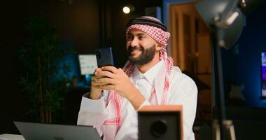 Portrait of happy arabic teleworker sending text messages to friends while working from home in stylish apartment. Cheerful Middle Eastern man typing on mobile phone with opened tv as background noise photo