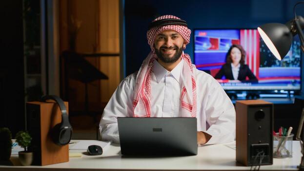 Portrait of smiling Arab businessman working at modern desk, typing on his laptop, solving tasks. Muslim guy browsing on digital device, doing email communication in professional office setting photo