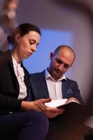 Businessman doing overtime with businesswoman late night afterhours tired on a deadline. Colleague entrepreneur working late together in evening at corporate job sitting on staircase, explaining difficult finance project. photo