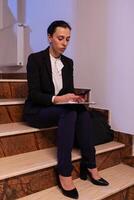 Exhausted tired businesswoman working on project deadline using smartphone. Serious entrepreneur working on job project sitting on staircase of business building at night hours for job. photo