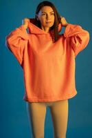 Young brunette in sports leggings and orange hooded top on blue background photo