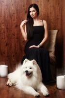 hermosa morena joven mujer largo negro vestir con un blanco como la nieve perro Samoyedo fornido estudio en sombras de marrón velas foto