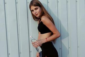 Female athlete drinking water while standing by a grey wall. photo