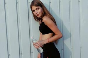 Female athlete drinking water while standing by a grey wall. photo