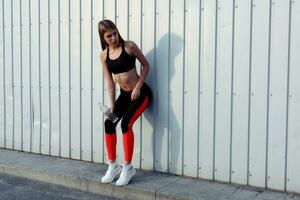 Female athlete drinking water while standing by a grey wall. photo