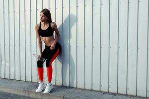 Female athlete drinking water while standing by a grey wall. photo