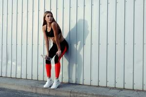 Female athlete drinking water while standing by a grey wall. photo