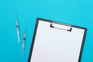 Vaccination, Immunology or Revaccination Concept Two Medical Syringe Lying on Blue Table in Doctor's Office in a Hospital or Clinic. Black Clipboard with Sheet of Paper Mock Up with Copy Space photo