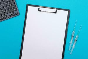 Vaccination, Immunology or Revaccination Concept Two Medical Syringe Lying on Blue Table in Doctor's Office in a Hospital or Clinic. Black Clipboard with Sheet of Paper Mock Up with Copy Space photo