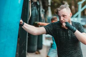 Street Fighter in Black Clothes and Bandages on the Wrist Boxing in Punching Bag Outdoors. Young Man Doing Box Training and Practicing His Punches at the Outside Gym photo