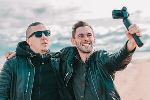Two Handsome Smiling Friends Making Selfie Using Action Camera with Gimbal Stabilizer at the Beach. Youthful Men in Black Clothes Having Fun by Making Photos