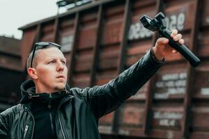 Handsome Guy in Black Clothes and Sunglasses Making Selfie or Streaming Video Using Action Camera with Gimbal Camera Stabilizer at Railway photo