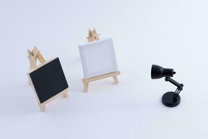 Two Wooden Easels Miniature with Blank Black and White Square Canvas for Artists and Painters Mockup. Mini Wooden Stand with Clean Artboard and Small Black Table Lamp on White Background, Copy Space photo