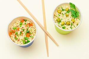 Prepared Instant Noodles with Green Pea, Carrot and Greens in Blue and Green Cups with Chopsticks on White Background. Quick Lunch or Unhealthy Fast Food photo