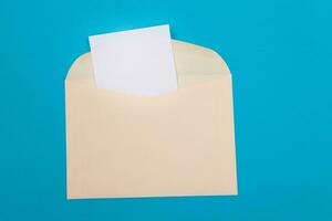 Beige Envelope with Blank White Sheet of Paper Inside, Lying on Blue Background Mock Up with Copy Space. Receiving Mail or Notice, Sending Postcard Top View, Flat Lay photo