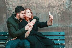 alegre emocional joven Pareja sentado en el banco y haciendo autofoto dos contento personas amor historia en el calle foto