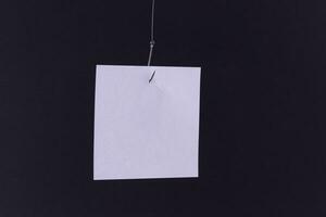 Mockup of a Blank White Memo Paper with Copy Space Hanging on a Fishing Hook Against the Black Background. Reminder or To Do List. Sticky Note Template photo