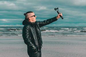 joven blogger en Gafas de sol haciendo selfie o transmisión vídeo a el playa utilizando acción cámara con cardán cámara estabilizador. hipster en negro ropa haciendo foto en contra el mar