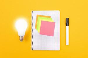 Glowing Power Saving Lamp and Notepad with Sticky Notes Lying on Yellow Table, Blank Template with Copy Space Top View, Flat Lay photo